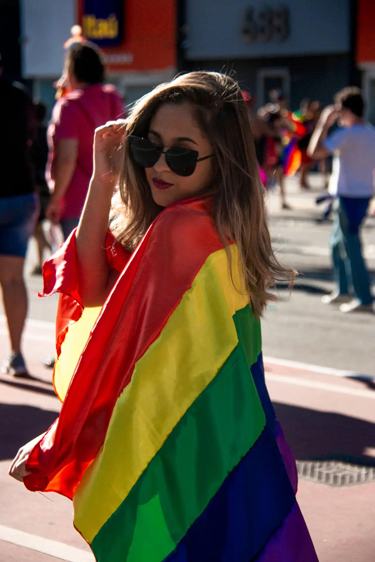 A smiling woman in sunglasses wrapped in a rainbow flag at a Pride parade.
