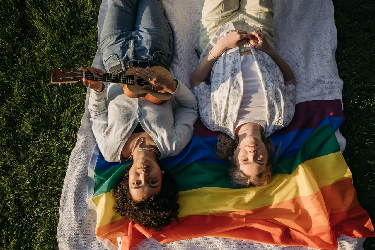 Friends lying on a rainbow blanket in the grass, one playing a ukulele.