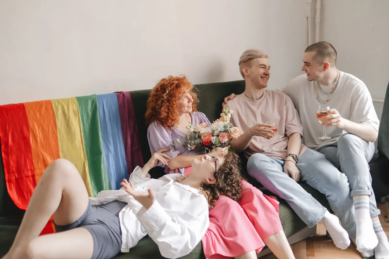 Friends laughing together on a sofa draped with a rainbow flag.